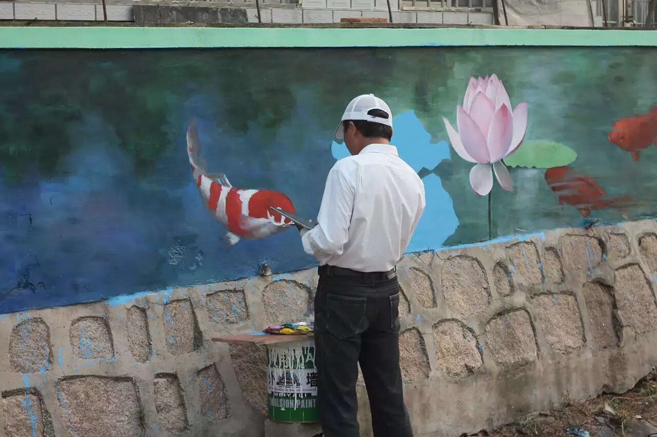 A painter seen from behind working on a koi and lotus mural above a stone-patterned wall.